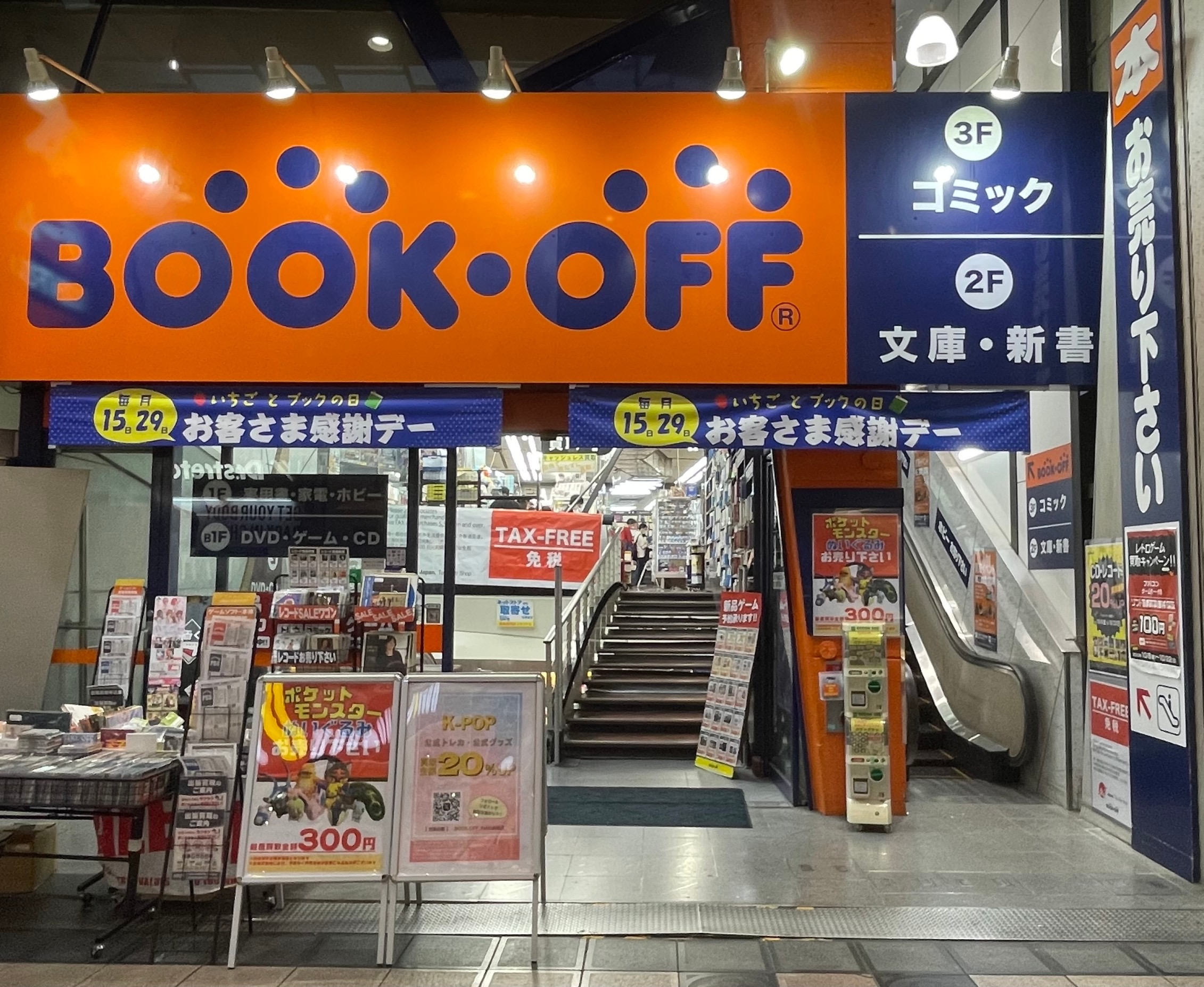 BOOK-OFF storefront with orange logo sign, blue floor guide for Gomic and books, promo banners, steps and escalator inside.